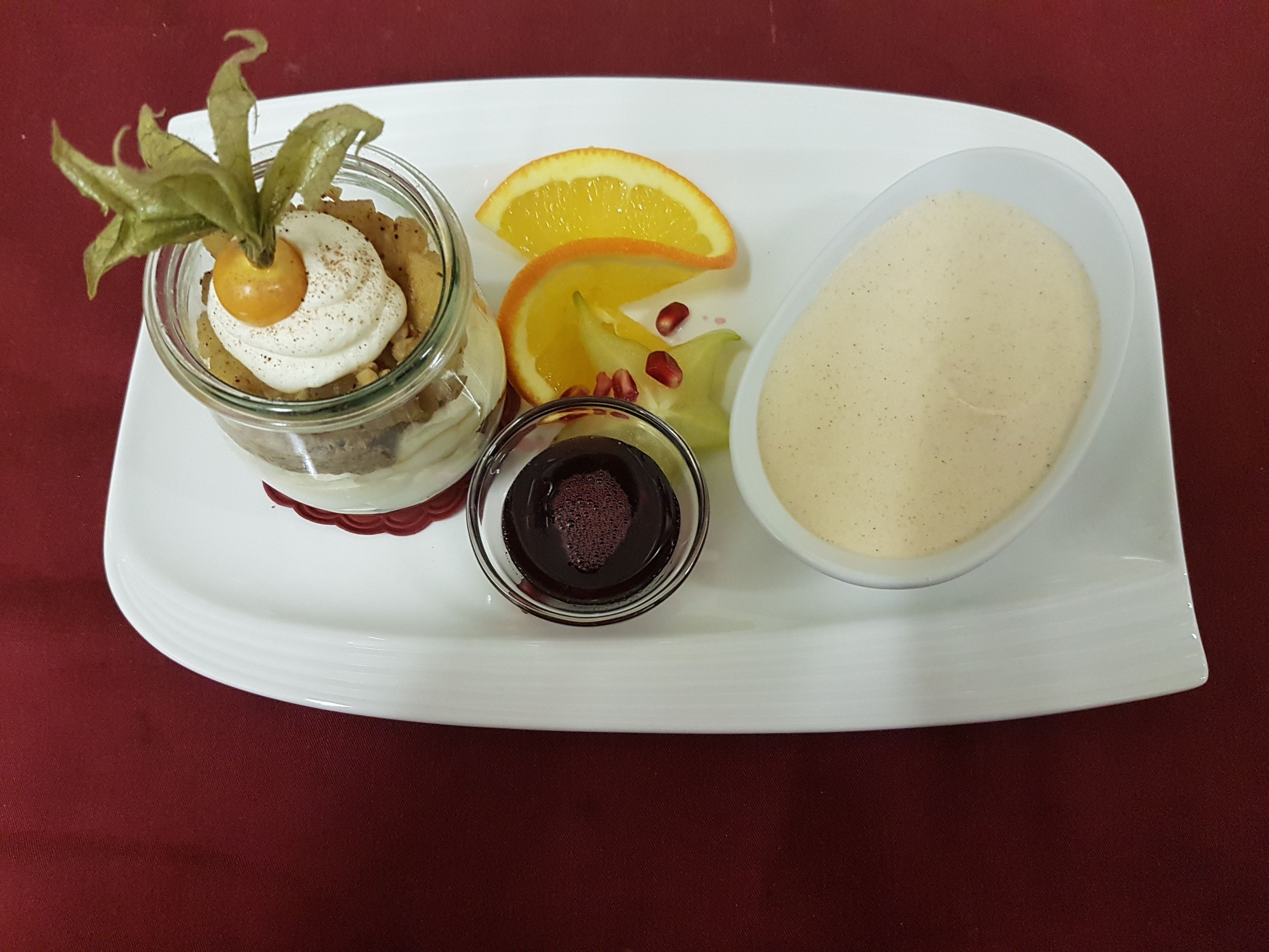 Ein Desserteller mit einem Glasgefäß, das ein mehrschichtiges Dessert enthält, einer Schüssel Sahne und zwei kleinen Schalen mit dunkler Flüssigkeit.
