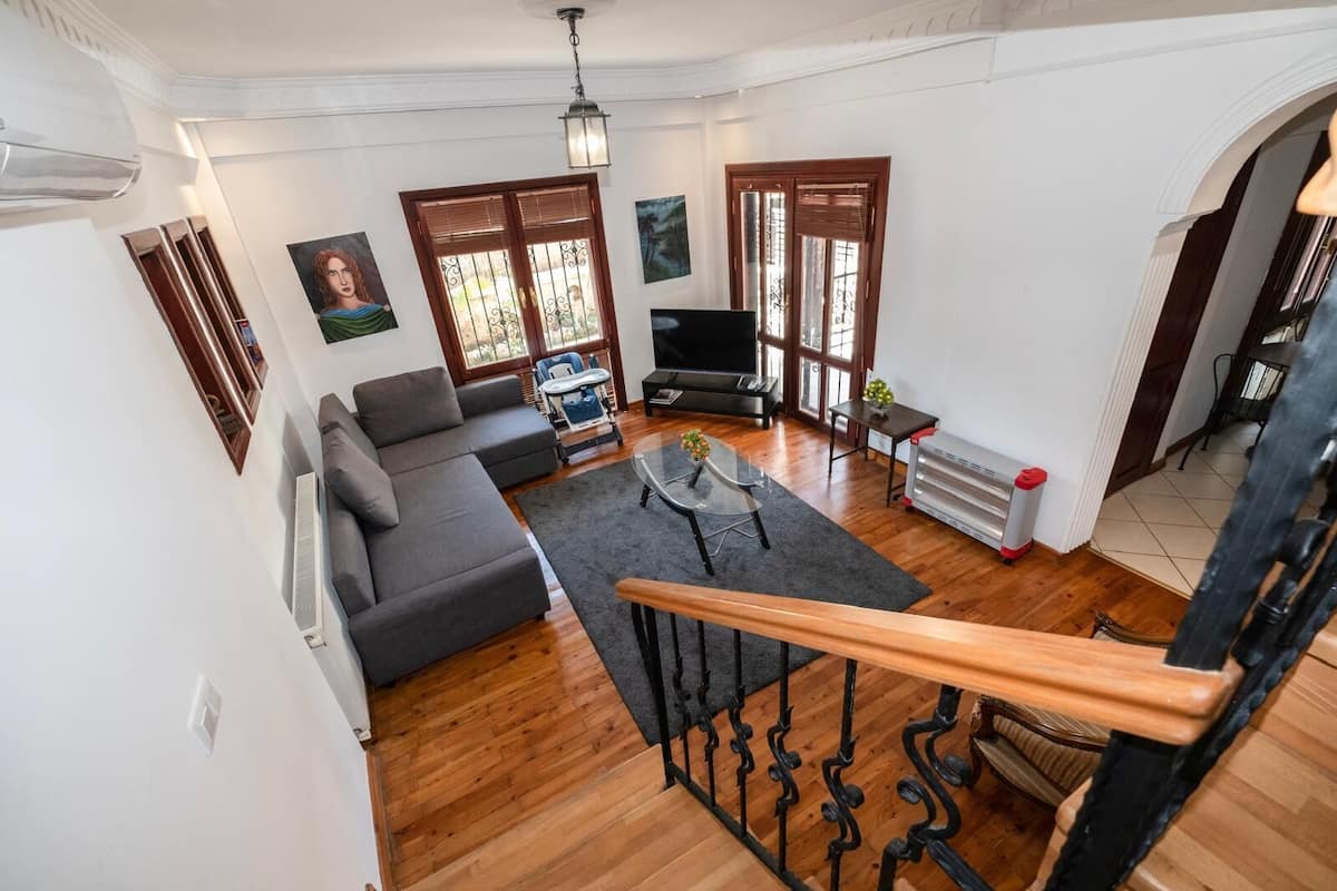 A spacious living room with a grey sofa, a coffee table, and a TV. There is a staircase with a wooden railing leading to another level.