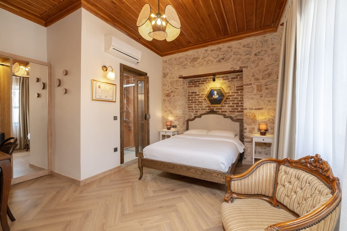 A bedroom with a wooden ceiling, a stone wall, a bed, bedside tables, and a chair.