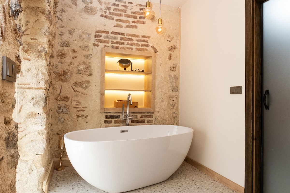 A modern bathroom with a freestanding bathtub, exposed brick walls, and a small shelf with lighting.