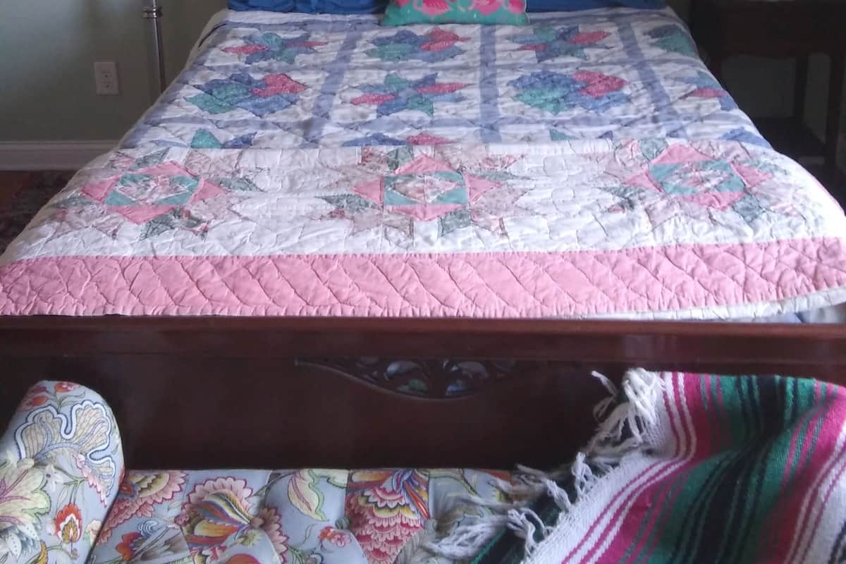 A bed with a colorful quilt, two pillows, a framed painting above, a lamp, and a bedside table with a lamp.