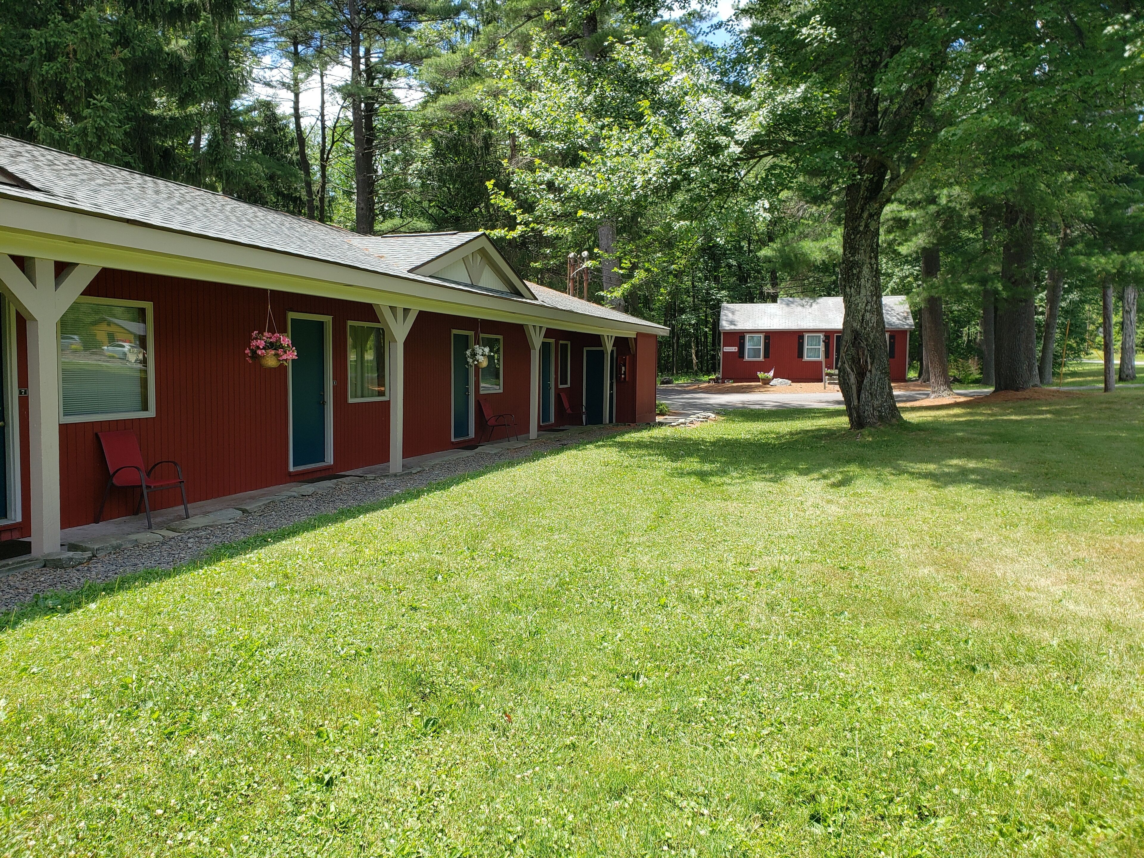 A row of red cabins with white trim, each with a door and a window, set in a grassy area with trees in the background.
