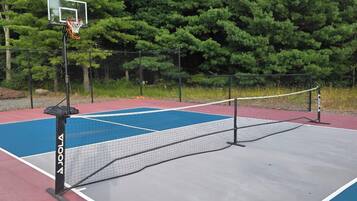 Cancha de tenis exterior con red, rodeada por una valla y árboles.