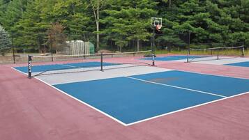 A tennis court with a net and a basketball hoop, surrounded by trees.