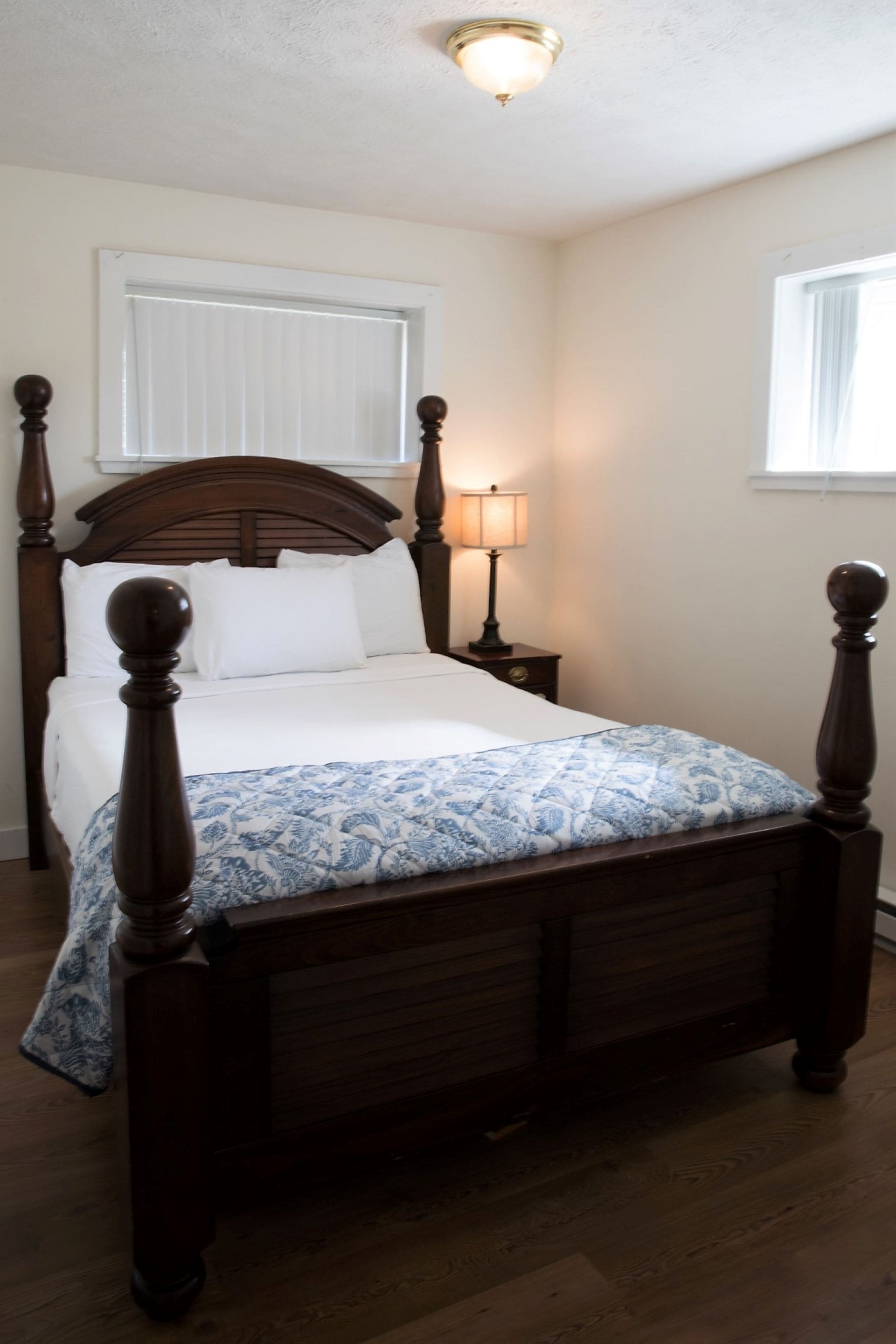 A wooden bed with a headboard, a bedside table with a lamp, and a window with blinds.