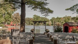 Un salón de bodas al aire libre con filas de sillas y mesas blancas, un arco de madera y un edificio rojo al fondo.