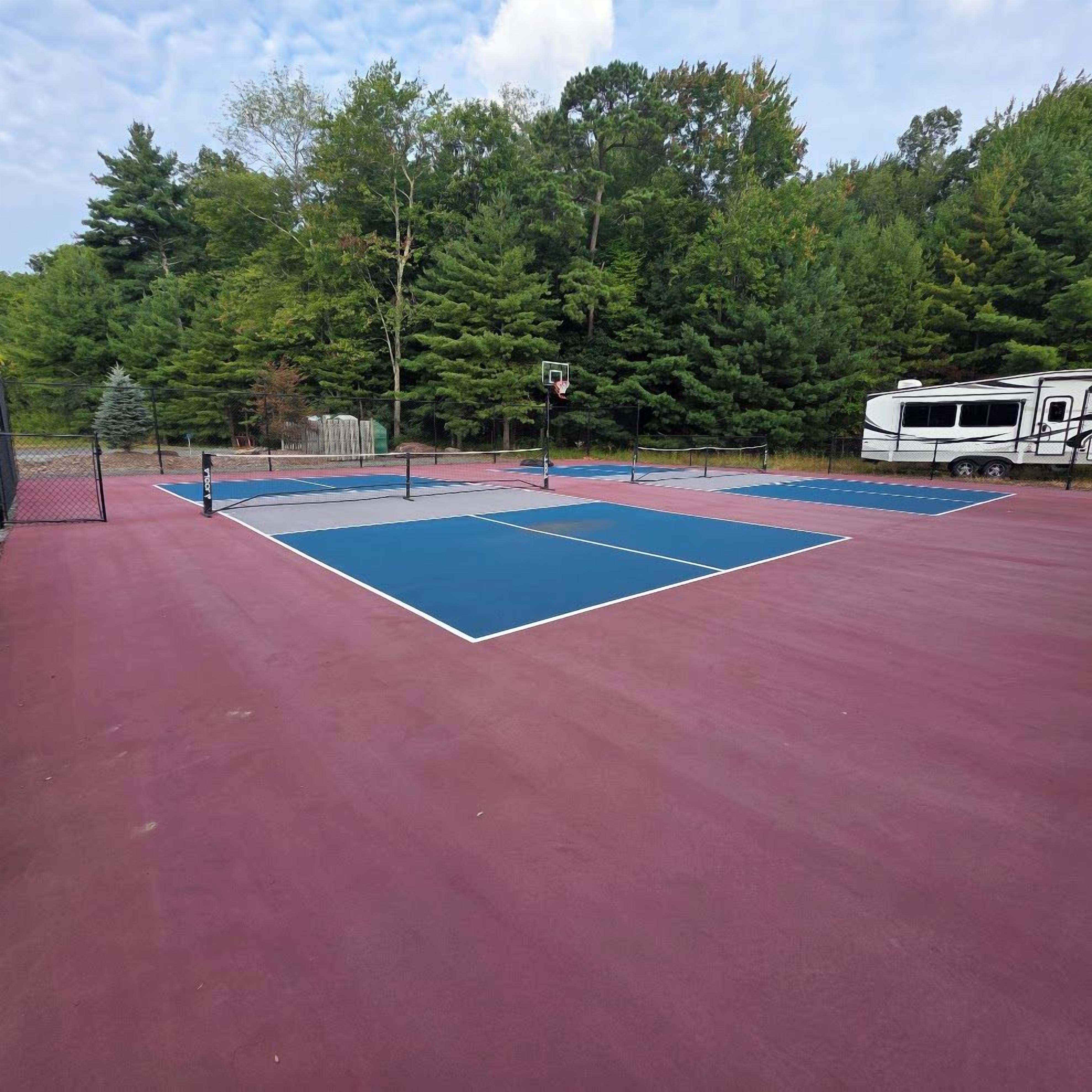 Un court de tennis extérieur doté d’une surface rouge et de zones de jeu bleues, entouré d’arbres et d’un camping-car blanc.