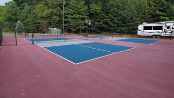 Un court de tennis extérieur doté d’une surface rouge et de zones de jeu bleues, entouré d’arbres et d’un camping-car blanc.