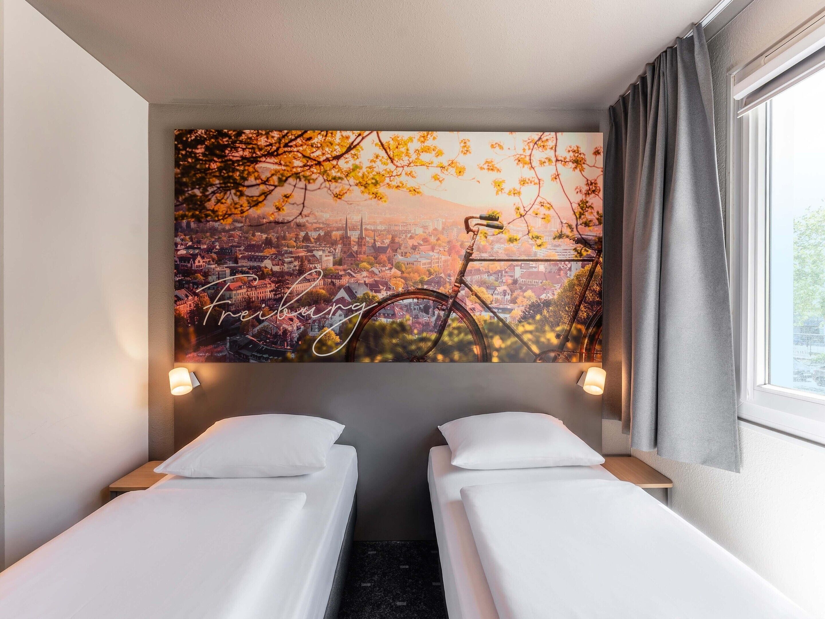 Habitación de hotel con dos camas individuales, un mural de paisaje urbano y una ventana con cortinas.