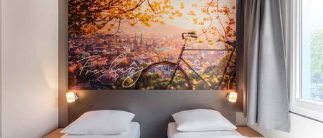 Habitación de hotel con dos camas individuales, un mural de paisaje urbano y una ventana con cortinas.