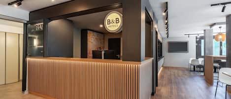 En receptionsdisk med et 'B&B' skilt, træpaneler og et moderne interiør med et fladskærms-tv og siddeområde.