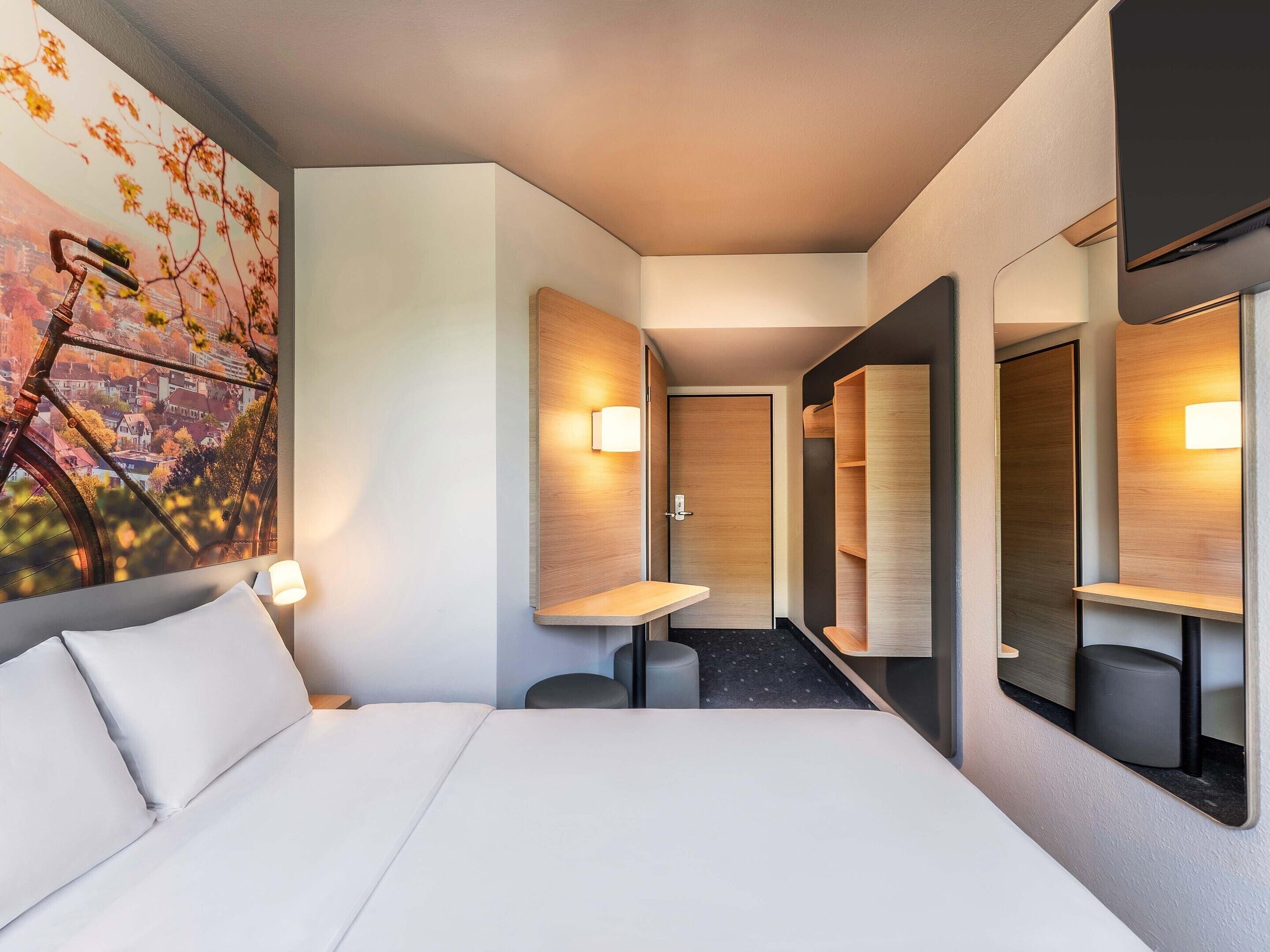 A hotel room with a bed, a painting of a bicycle, a mirror, and a small table.