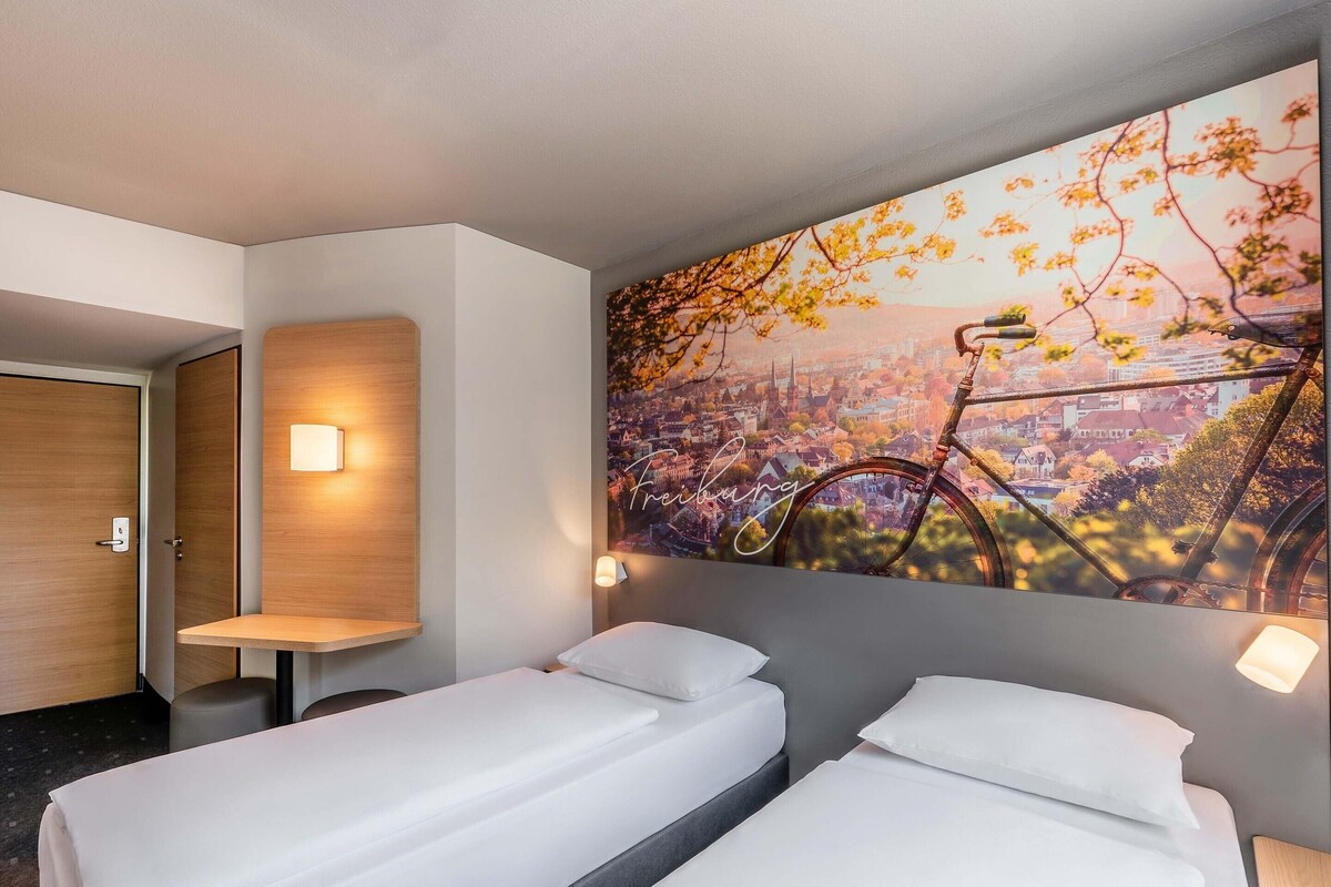Habitación de hotel con dos camas individuales, una puerta de madera, una mesita pequeña y un cuadro grande con una vista urbana.