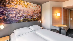 Una habitació d'hotel amb un llit, una taula de nit, una làmpada penjada a la paret i un ampli quadre amb una vista urbana.