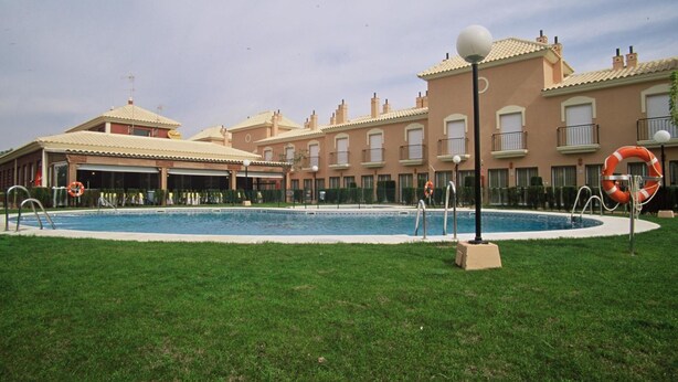 A swimming pool surrounded by a grassy area, with a building featuring balconies and a tiled roof in the background. - Interpass Golf Playa Country Club (Isla Cristina)