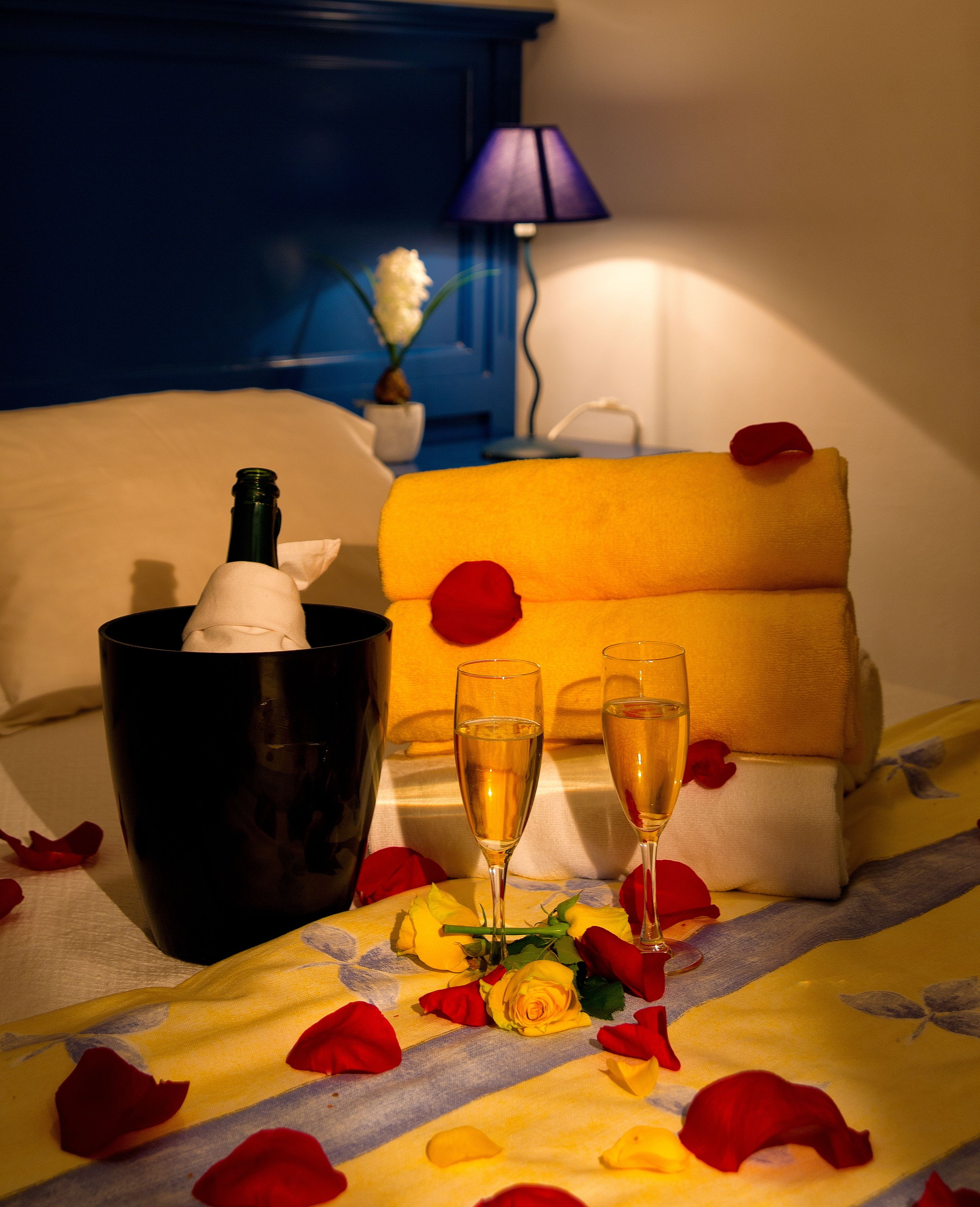 Un lit recouvert d’une couverture jaune, deux coupes à champagne, une bouteille de champagne et des pétales de rose éparpillés.