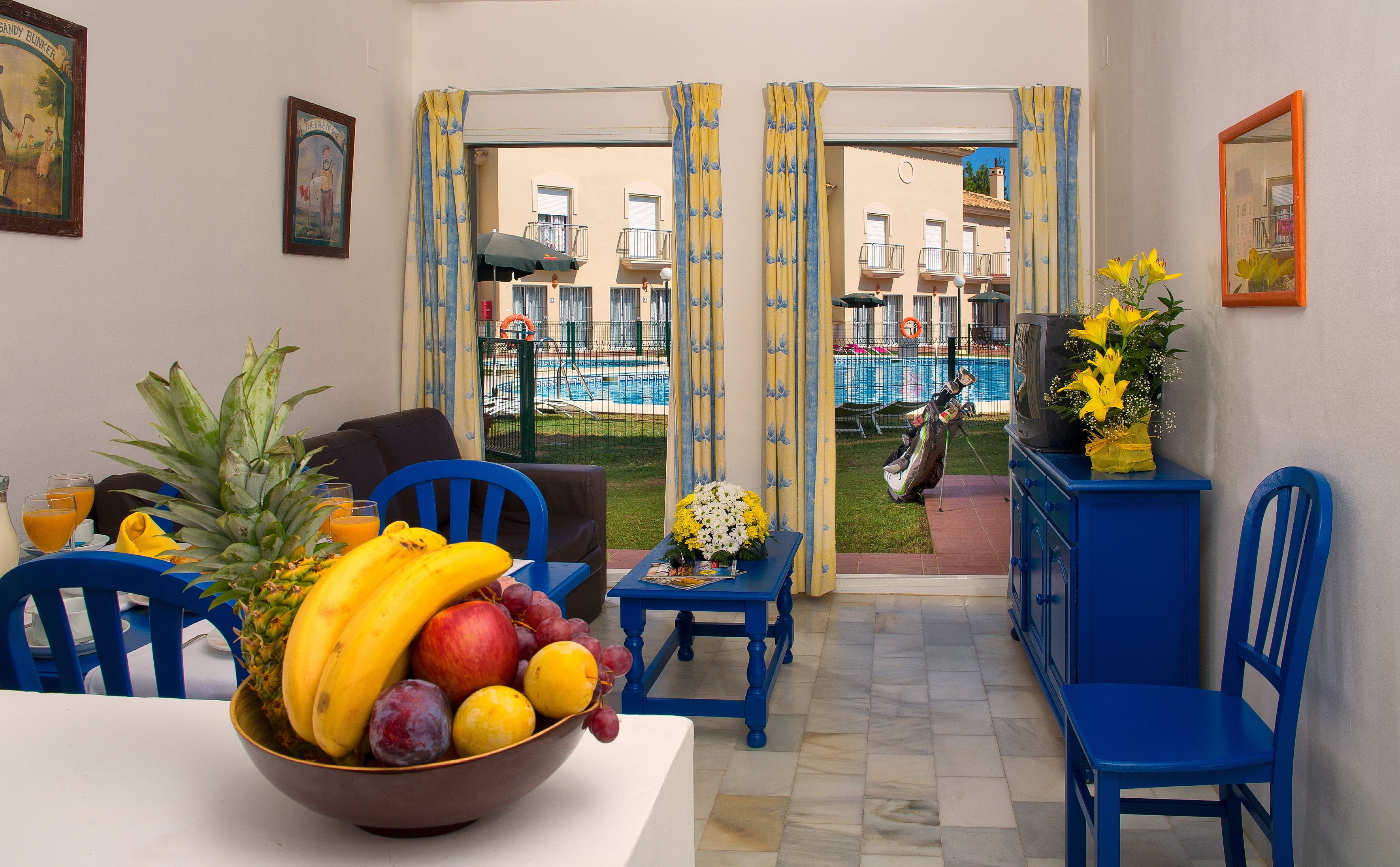 Un comedor con frutero, sillas azules, una mesa y vista a una piscina y edificios al fondo.