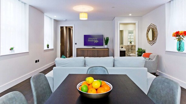 Un soggiorno moderno con un divano, un tavolo da pranzo con una ciotola di frutta, una televisione e un bagno visibile attraverso una porta aperta.