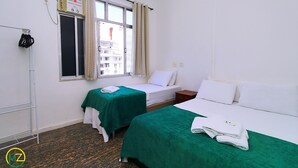 Ein Hotelzimmer mit zwei Betten, einem Fenster mit Vorhängen, einem Heizkörper und einem Regal mit Handtüchern.