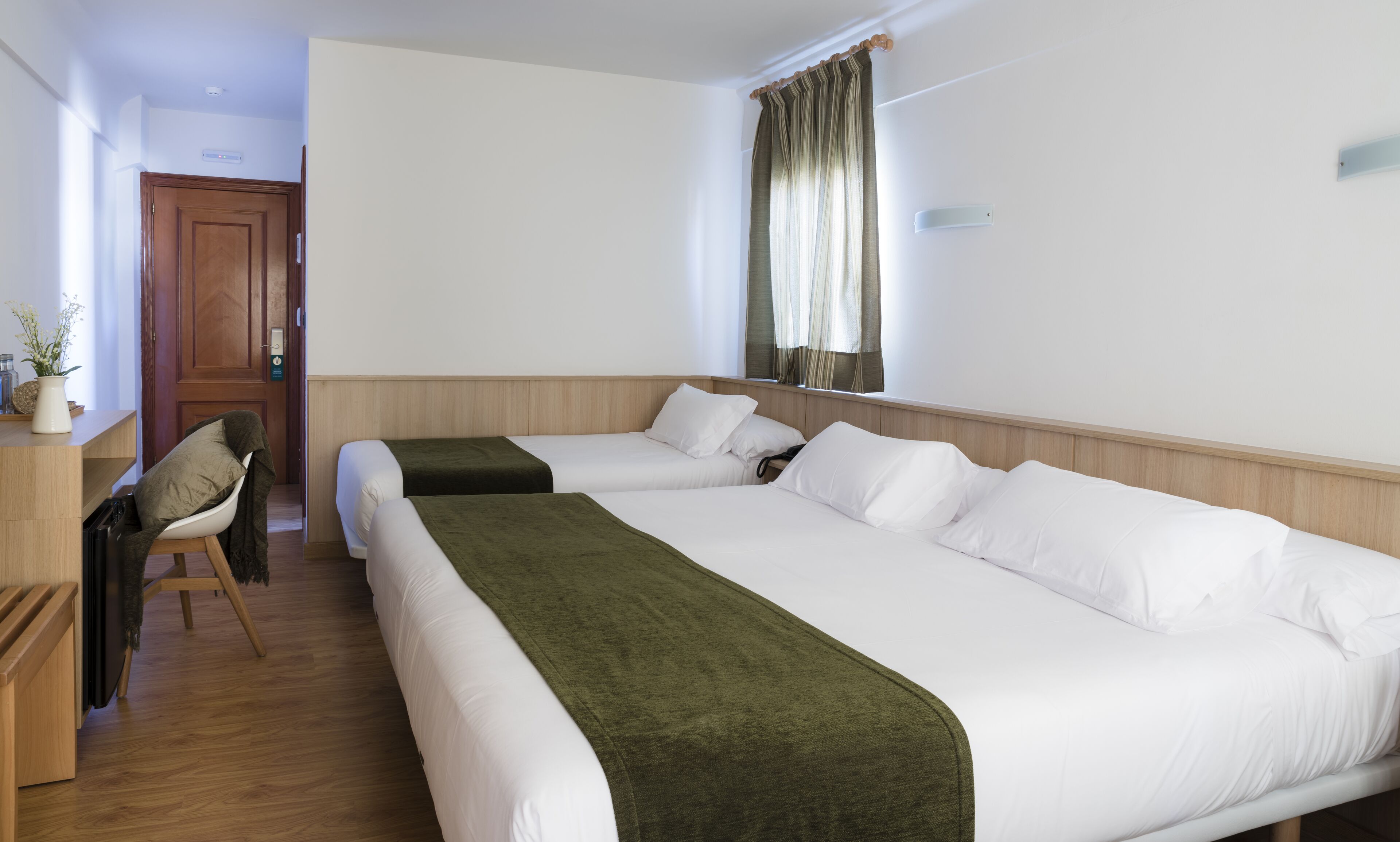 Une chambre d’hôtel avec deux lits simples, un bureau en bois, une chaise, une petite table avec un vase et une fenêtre avec des rideaux.