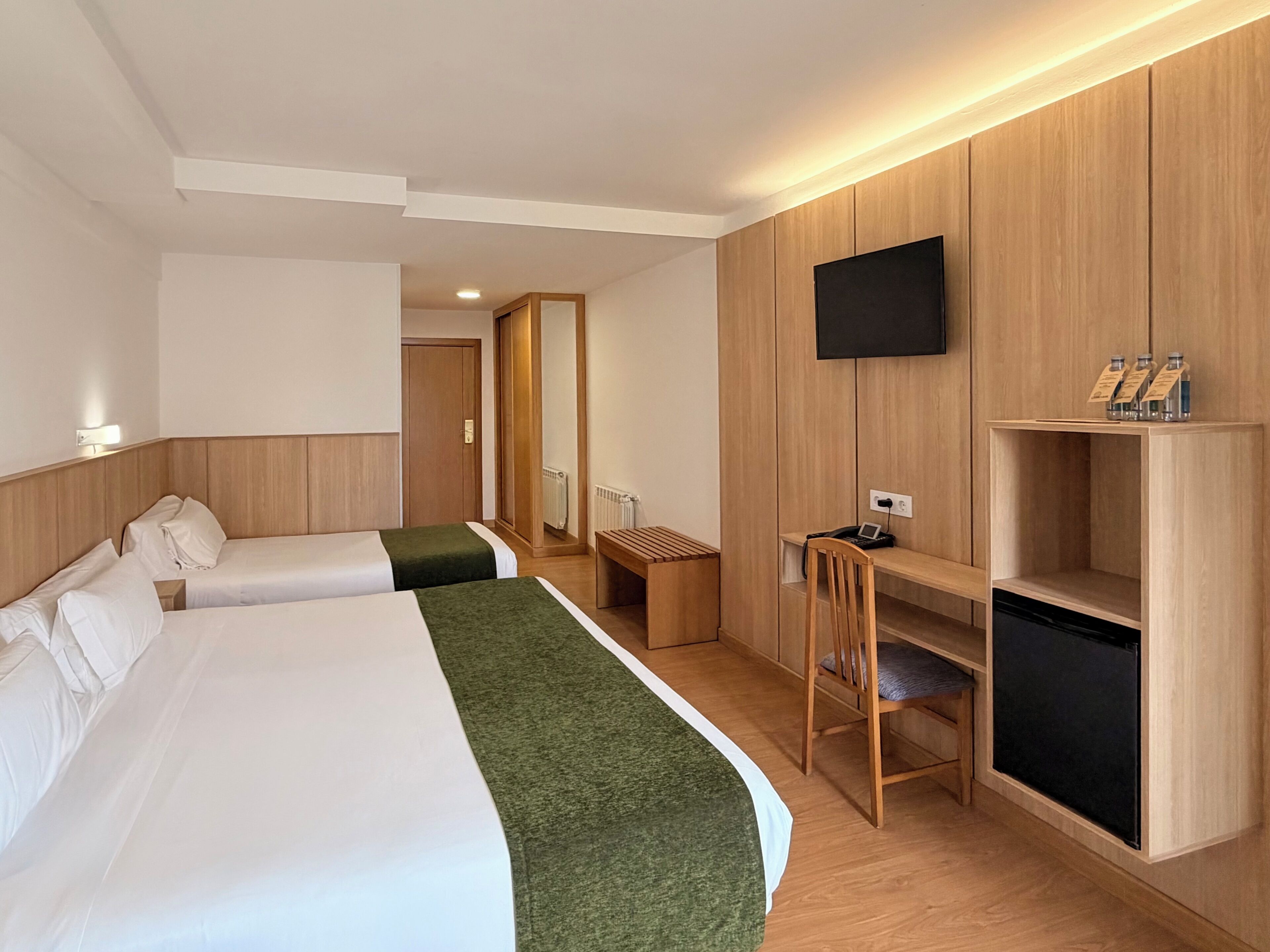 A hotel room with two beds, a desk, a chair, a TV, and a shelf with bottles.