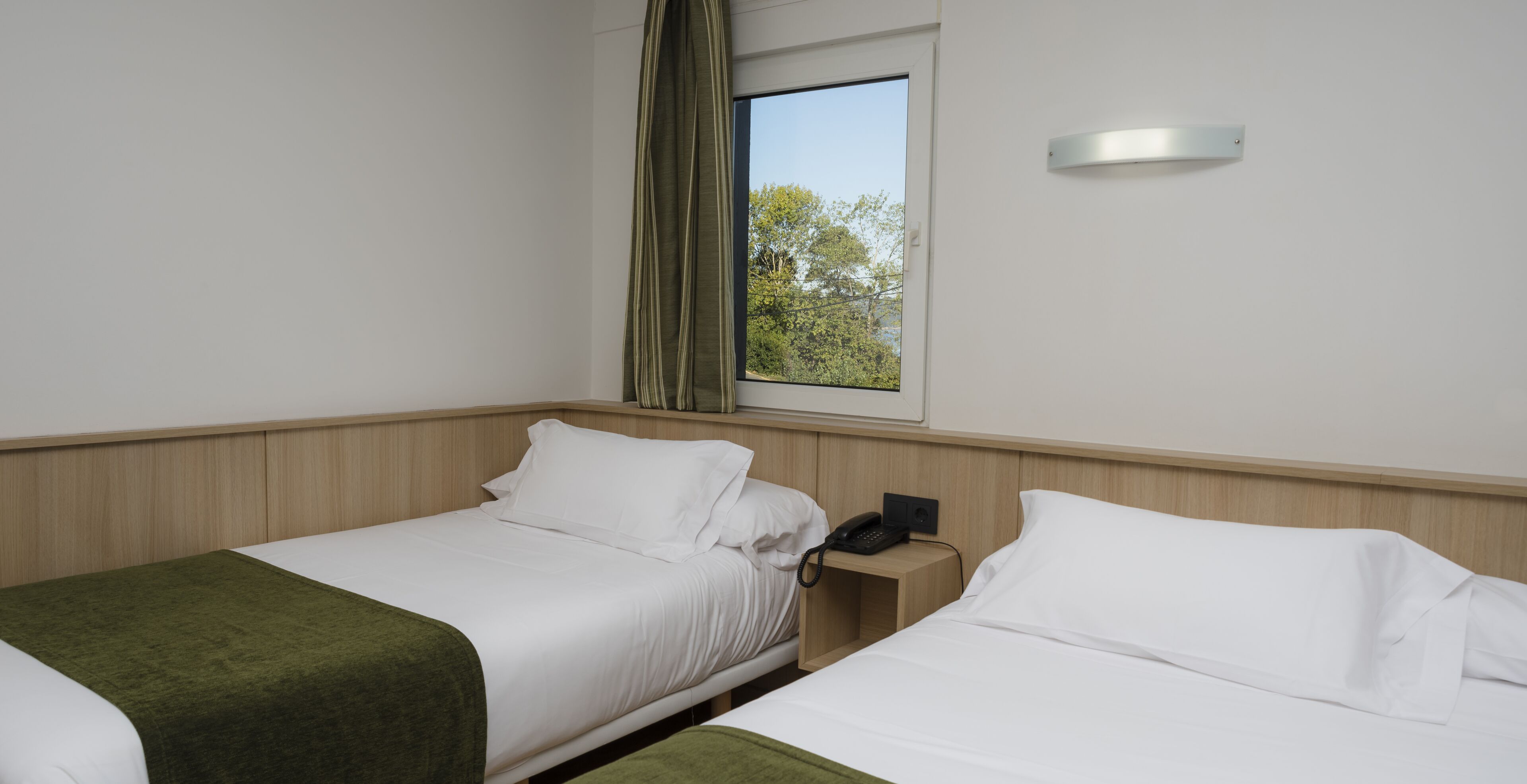A hotel room with two beds, a window with a view of trees, a bedside table with a phone, and a light switch on the wall.