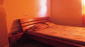 Une petite chambre à coucher peu éclairée, avec un lit simple, une table de chevet et une prise électrique au mur.