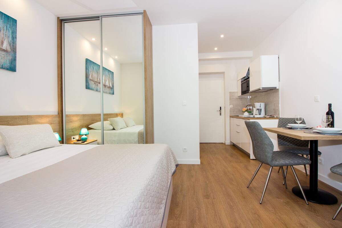 A hotel room with a bed, a desk, a chair, a mirror, and a kitchen area.