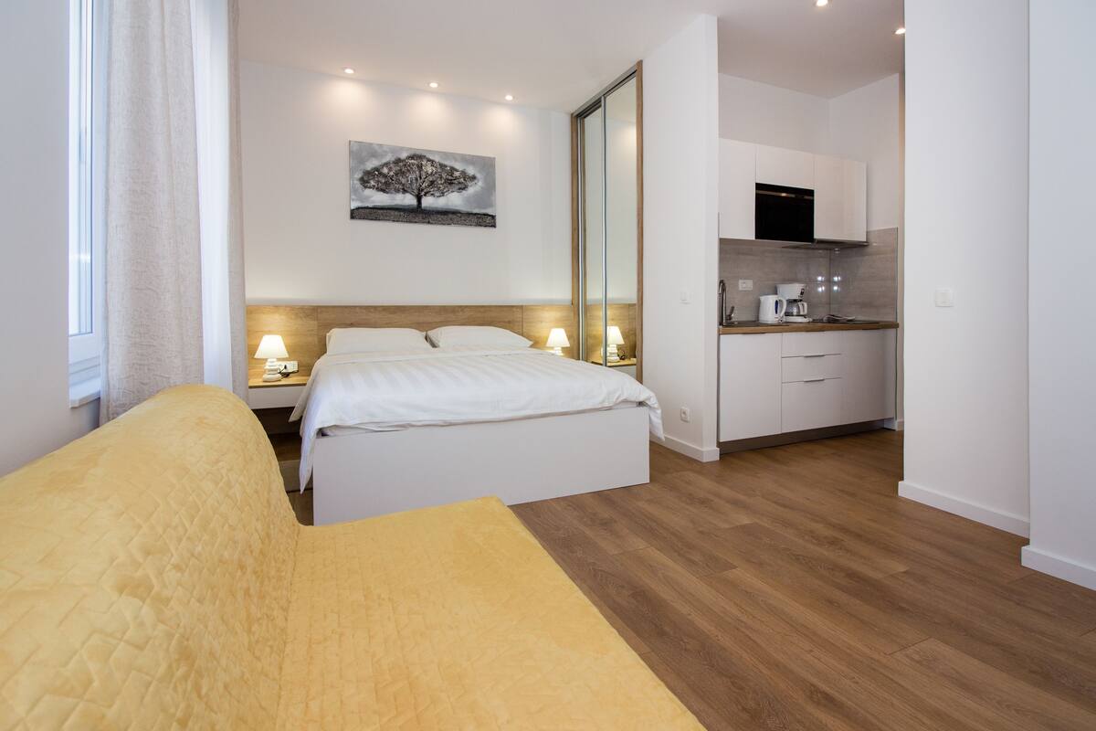 A modern hotel room with a bed, bedside lamps, a painting, a kitchen area, and wooden flooring.