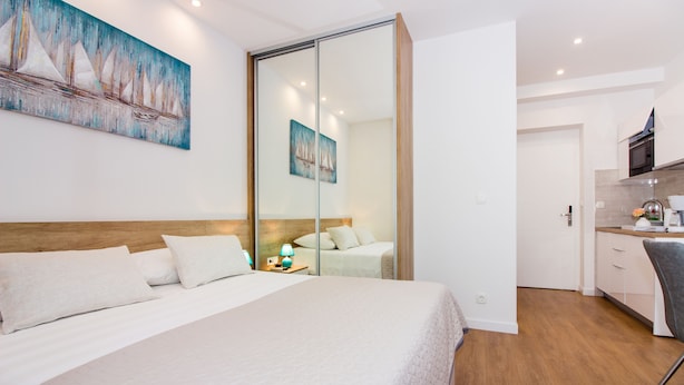 A hotel room with a large bed, a wooden headboard, a nightstand with a lamp, and a sliding door leading to a kitchen area. - Lugo Apartments (Split)