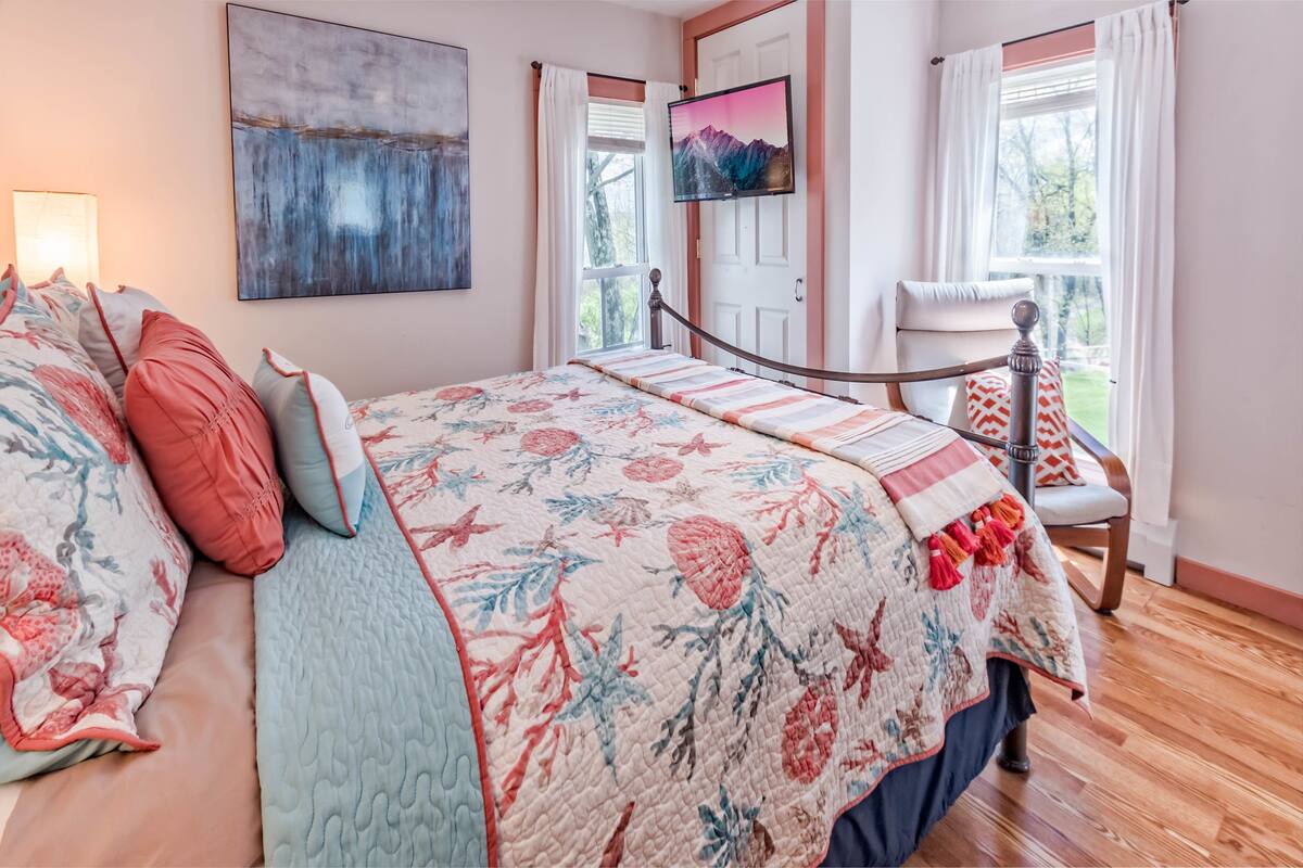 A bedroom with a floral bedspread, a mounted TV, a wooden chair, and a view of the outdoors through a window.