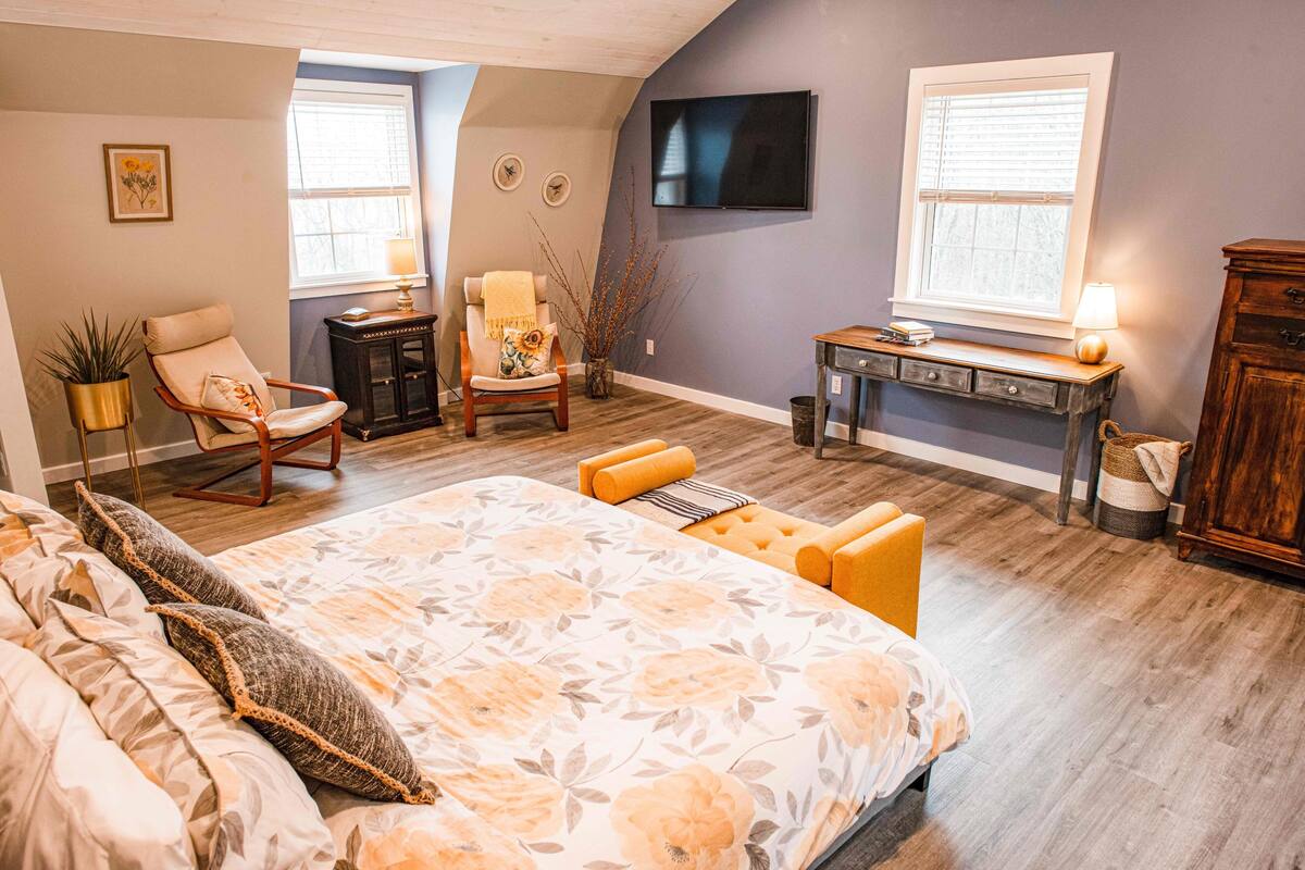 A bedroom with a large bed, two chairs, a desk, and a TV on the wall.