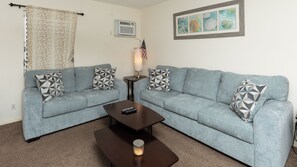 Ein Wohnzimmer mit zwei hellblauen Sofas, gemusterten Kissen, einem kleinen Tisch mit einer Fernbedienung, einer Lampe und einem gerahmten Bild an der Wand.