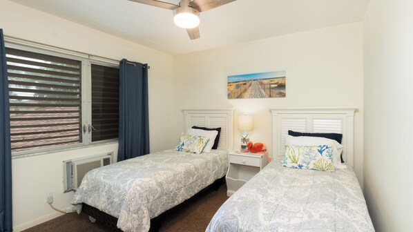 A bedroom with two beds, a ceiling fan, a window with blinds, a radiator, and a painting on the wall.