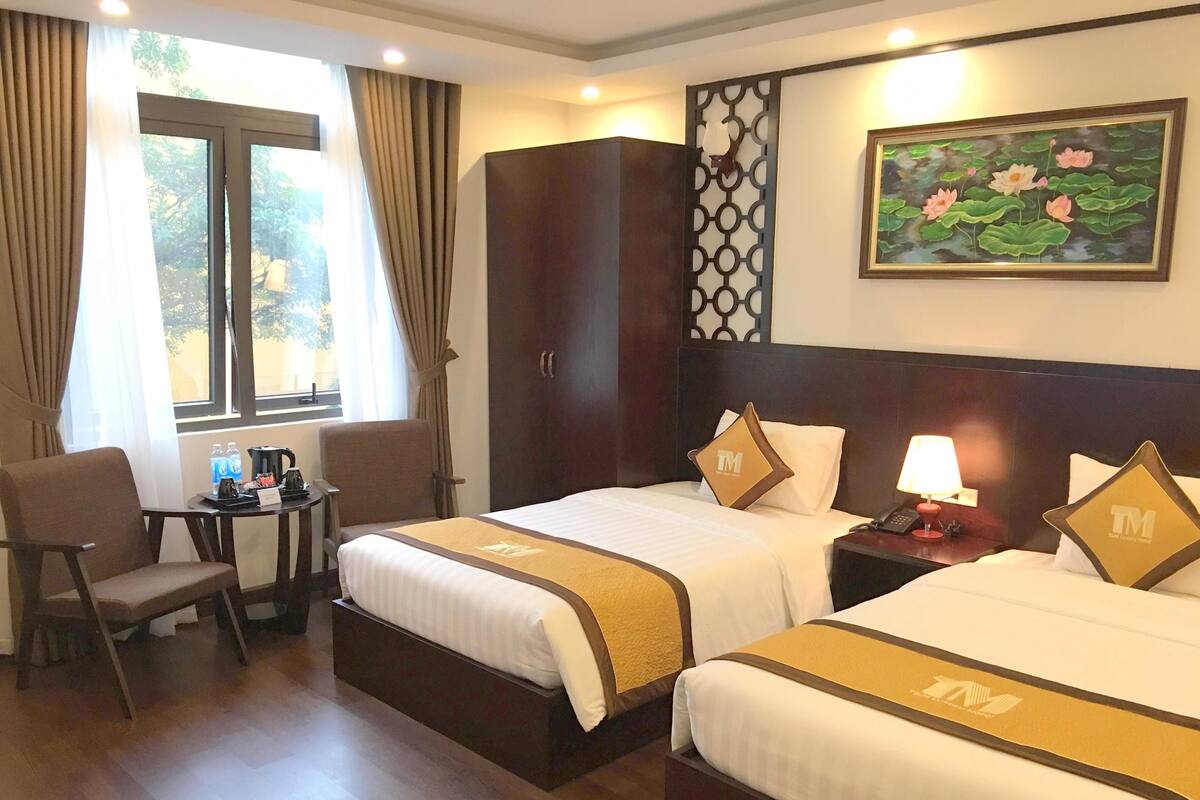 A hotel room with two beds, a desk with a chair, a wardrobe, and a window with curtains.