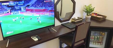 Une chambre d’hôtel avec une télévision posée sur un bureau en bois, une chaise, un petit réfrigérateur, un miroir et une poubelle.