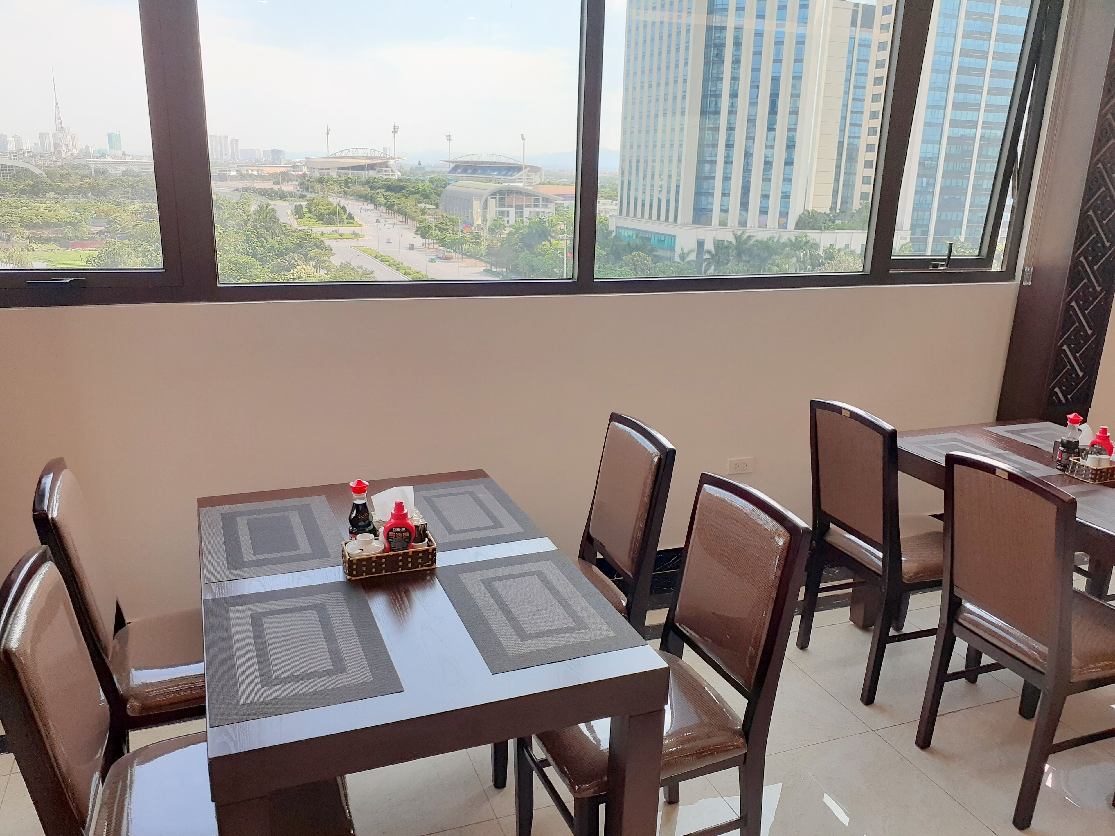 Un espace repas avec des tables et des chaises, une vue sur le paysage urbain à travers de grandes fenêtres, et une petite table avec des condiments.