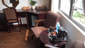 Quarto de hotel com escrivaninha de madeira, uma cadeira, uma mesinha com bandeja de itens de café da manhã, uma planta em vaso e uma janela com vista para o verde.