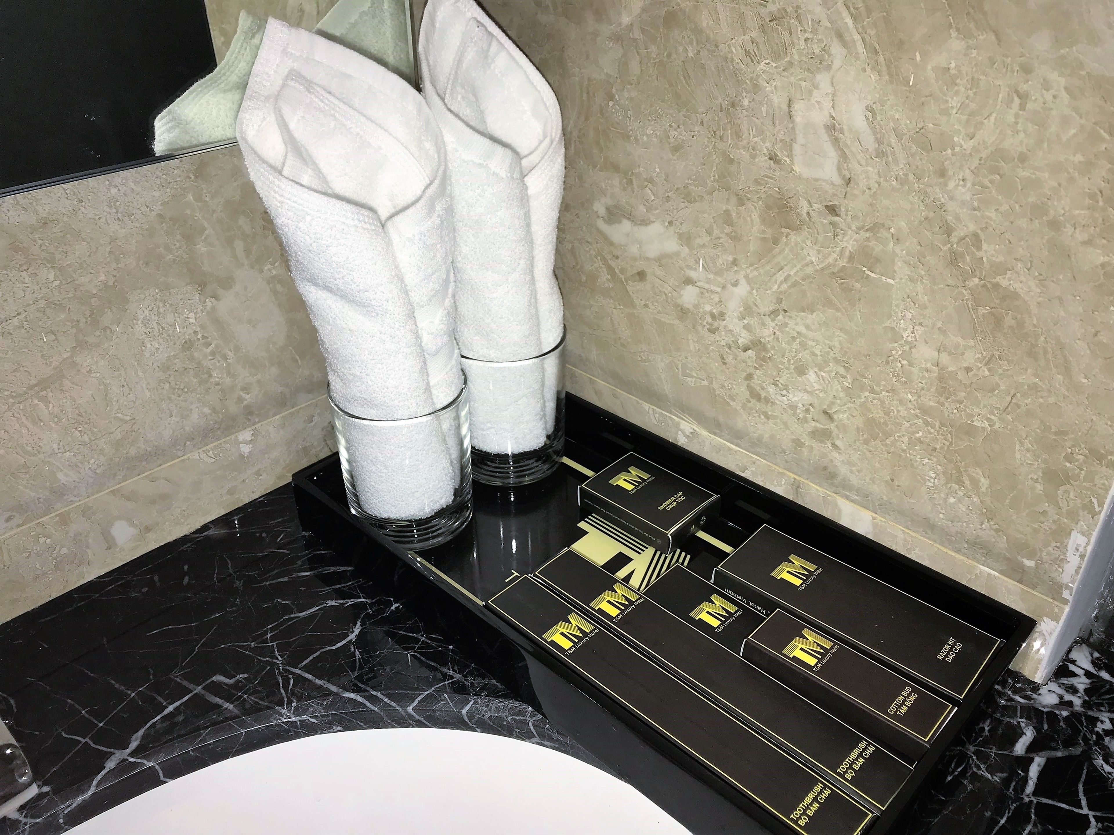 A bathroom with a black marble countertop, a white sink, and a towel rack with two white towels. There are black boxes with gold lettering on the countertop.