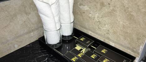 Un baño con encimera de mármol negro, un lavamanos blanco y un perchero con dos toallas blancas. Hay cajas negras con letras doradas sobre la encimera.