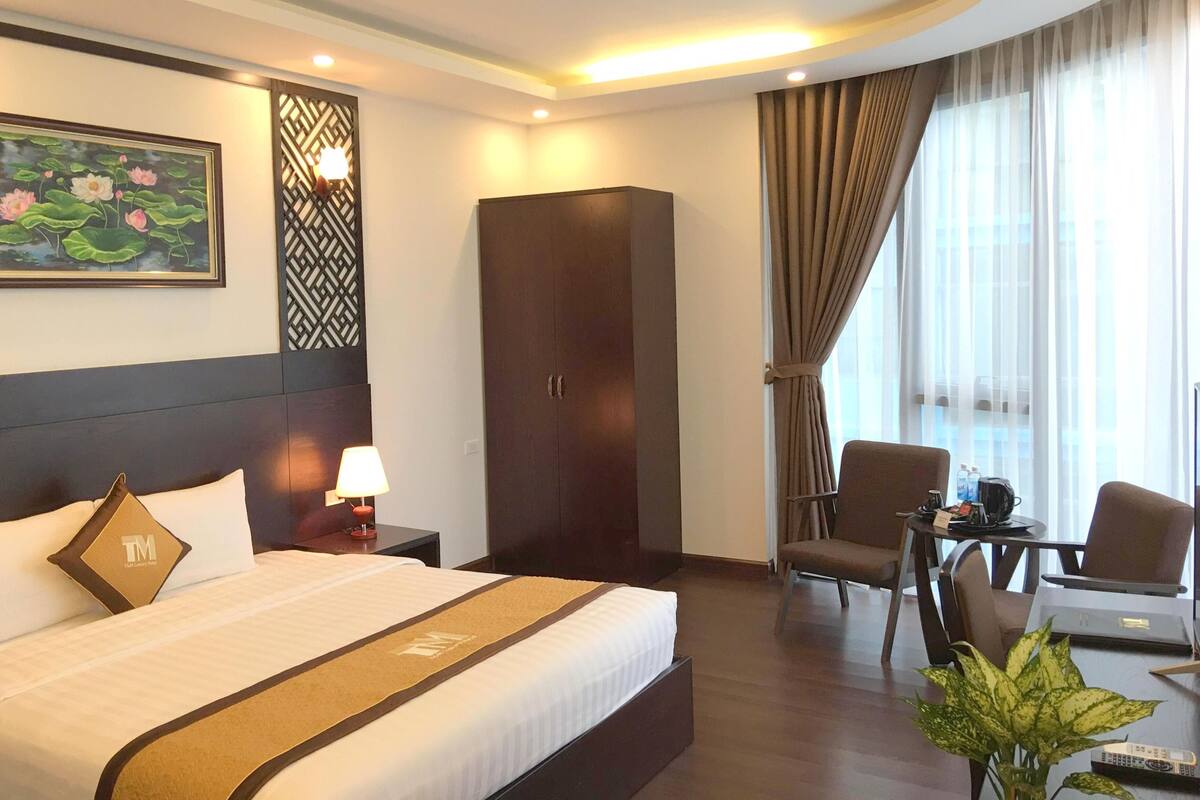 A hotel room with a large bed, a wardrobe, a desk with chairs, a painting of lotus flowers, and a window with curtains.