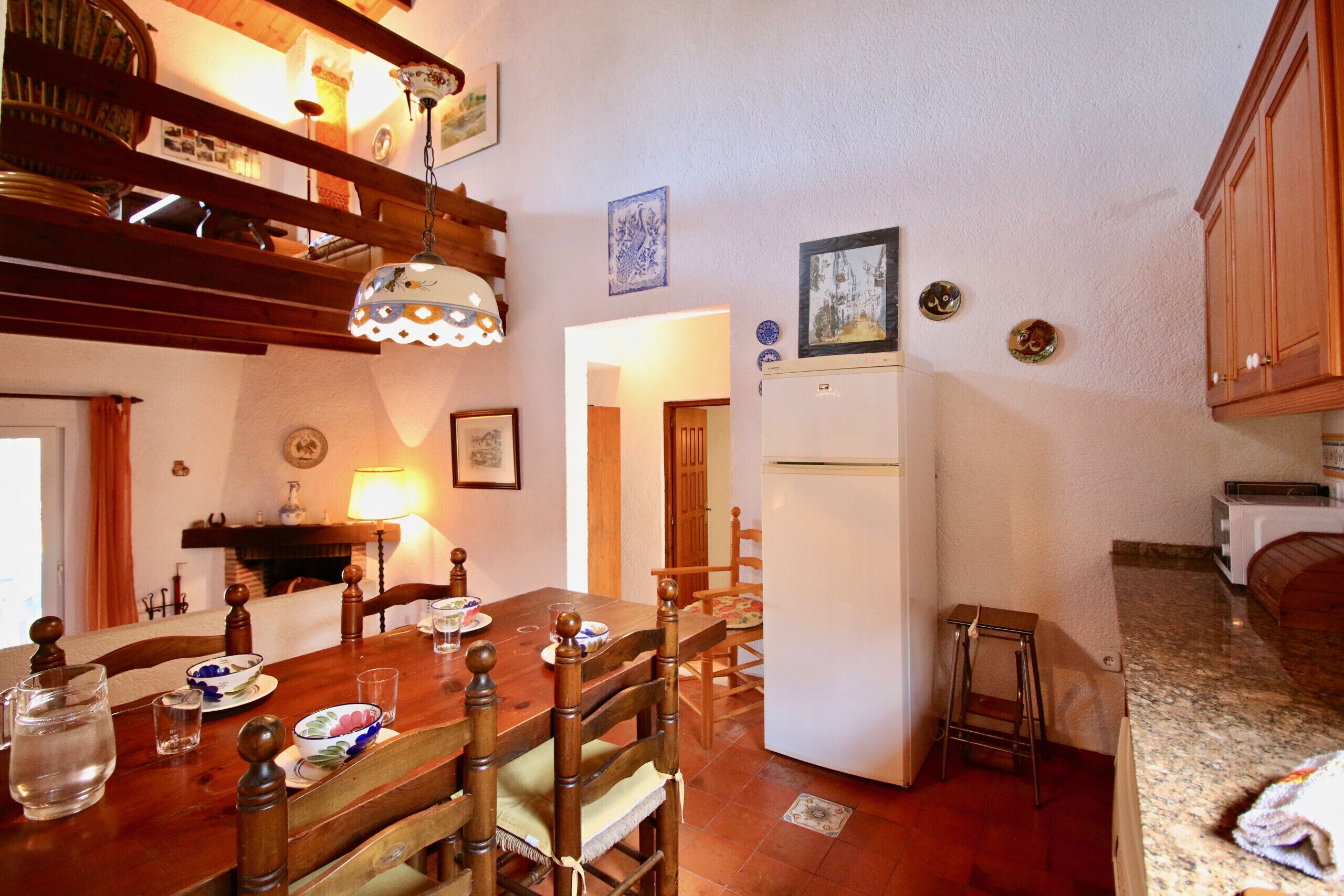 A dining area with a wooden table, chairs, a hanging light fixture, and a refrigerator.