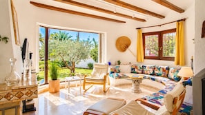 A sunlit living room with a sofa, chairs, a coffee table, and a view of an outdoor garden. - Villa Bellavista WA with Private Pool and Garden in Dénia (Denia)