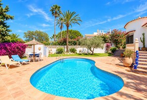 A swimming pool with a curved edge, surrounded by lounge chairs and a table, with palm trees and a white building in the background. - Villa Bellavista WA with Private Pool and Garden in Dénia (Denia)