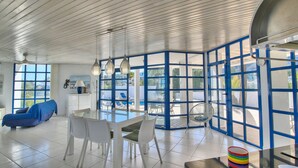 Une pièce spacieuse avec un plafond haut, un coin repas avec des chaises blanches et une table en verre, un canapé bleu et de grandes fenêtres aux montures bleues.
