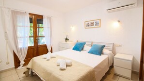 A bedroom with a bed, white curtains, a nightstand, a bedside table, a wall-mounted air conditioner, and a framed picture.