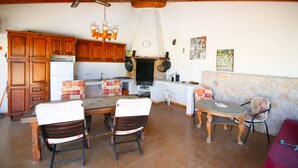 A rustic kitchen with wooden cabinets, a built-in stove, and a stone wall. There are two dining tables with chairs, one with a red tablecloth.