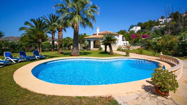 A swimming pool with a tiled edge, surrounded by palm trees and a white building with a tiled roof.