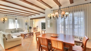 Uma sala de estar ampla com sofá, mesa de jantar e cadeiras. Há uma televisão ao fundo, e o teto possui vigas de madeira.