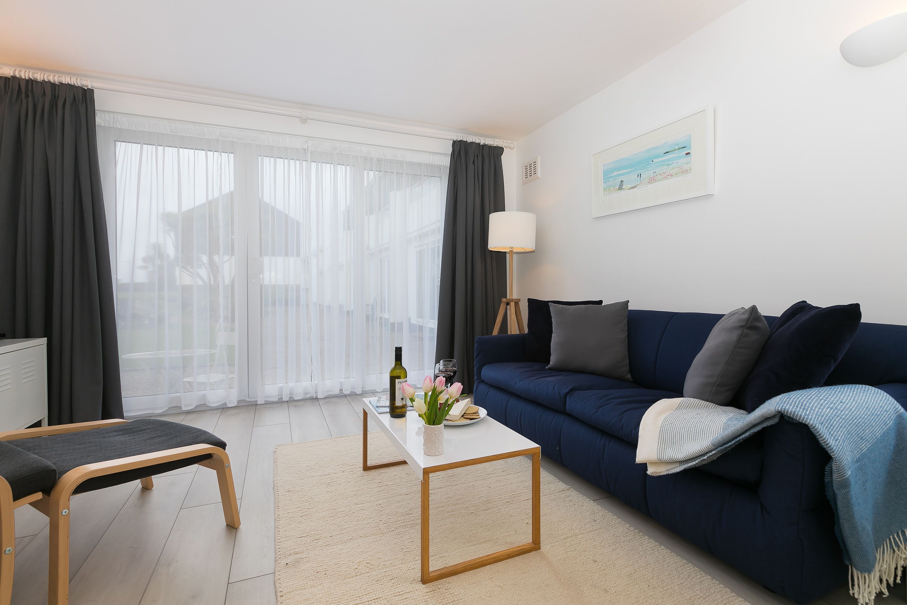 Un salon moderne avec un canapé bleu foncé, une table basse blanche et un banc. On y trouve une lampe, un tableau au mur et une fenêtre avec des rideaux légers.